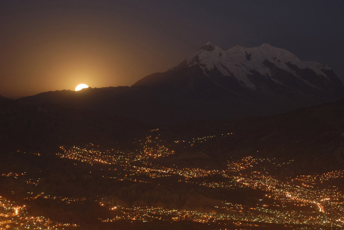 8.9.06 | A full moon rises over the city lights of La Paz.  The coppery color of the moonrise is a result of smog and dust in the atmosphere.  Illimani, the highest mountain in Bolivia at 21,125/6,439m, rises above the city.Every glacier in the central and northern Andes, has been in rapid retreat since the late 1970âs. The lower glaciers are thinning at the average rate of 1.1 meters per year; the higher glaciers at 0.5 meters per year. With the human populations below the peaks dependant on the water stored in fast-shrinking glaciers like this one, trouble lies ahead if these melting trends continue.